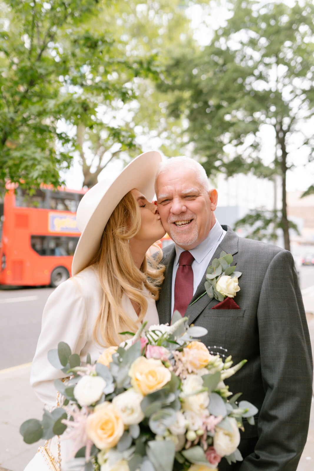 A woman in a white outfit and hat kisses an older man on the cheek while holding a bouquet of flowers. They are smiling outdoors, with a red double-decker bus and trees in the background.