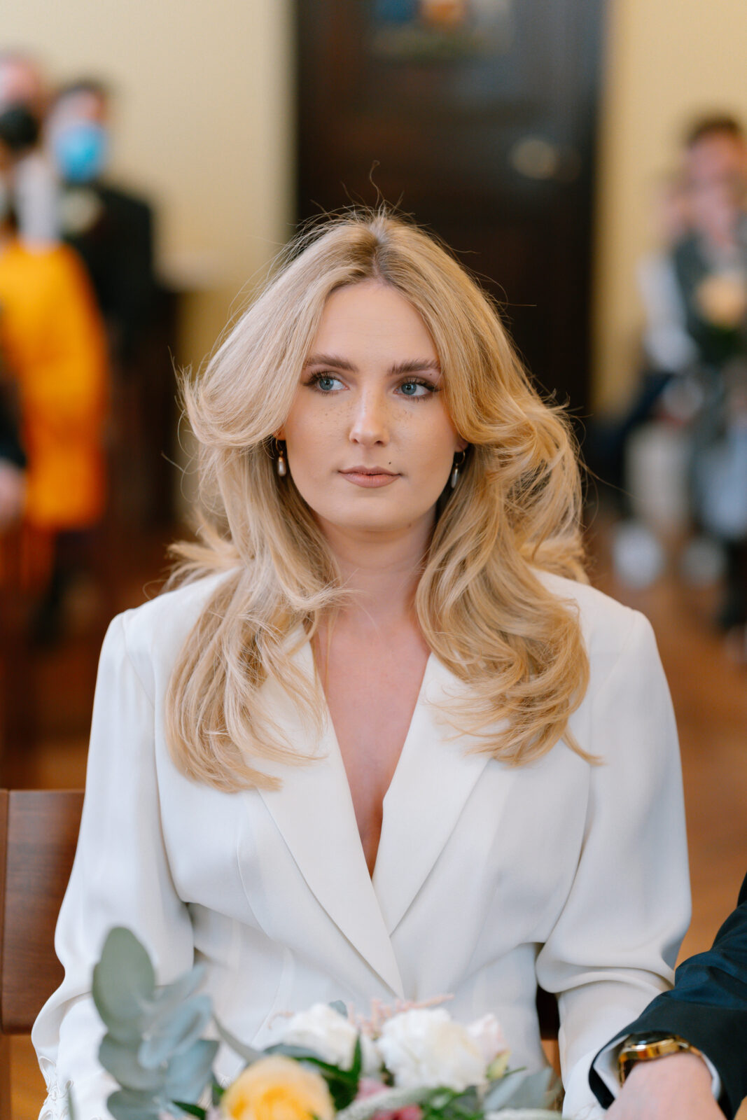 A woman with long, wavy blonde hair wearing a white suit sits indoors, holding a bouquet of flowers. She looks slightly to the side with a calm expression. People and blurred details are visible in the background.