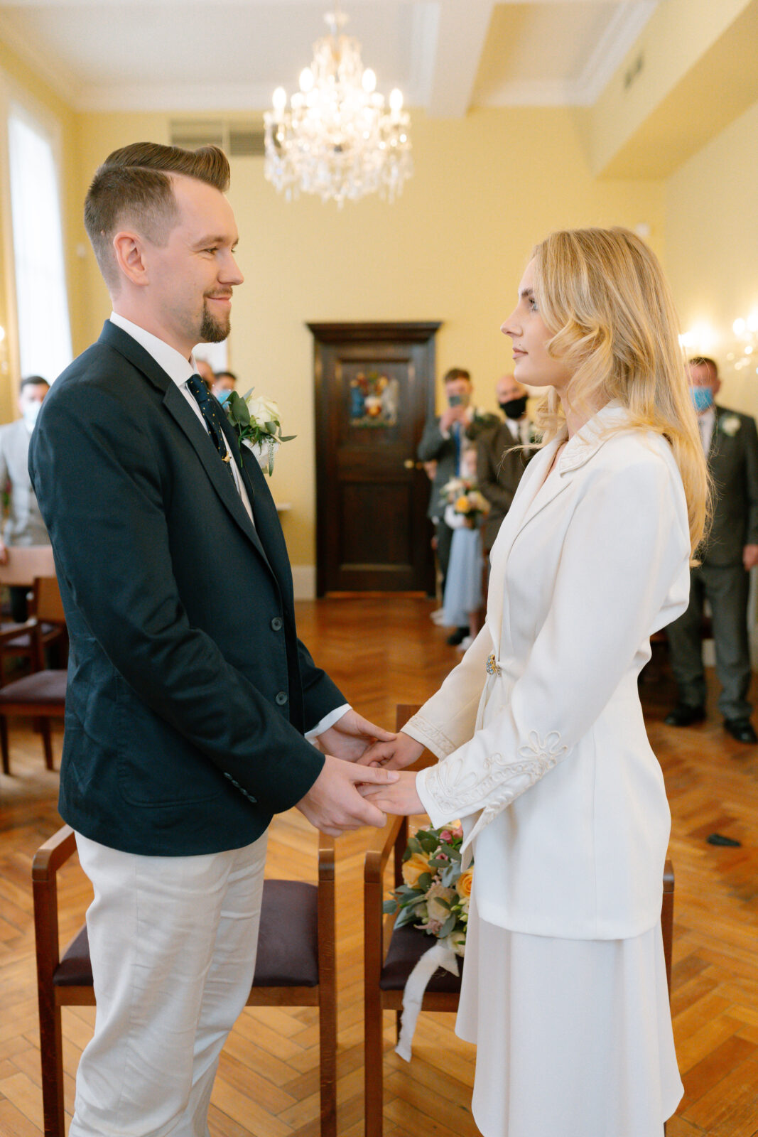 A man and woman stand facing each other, holding hands, in a formal room with chandeliers. They are dressed in semi-formal attire, suggesting a wedding or civil ceremony, with several people watching in the background.