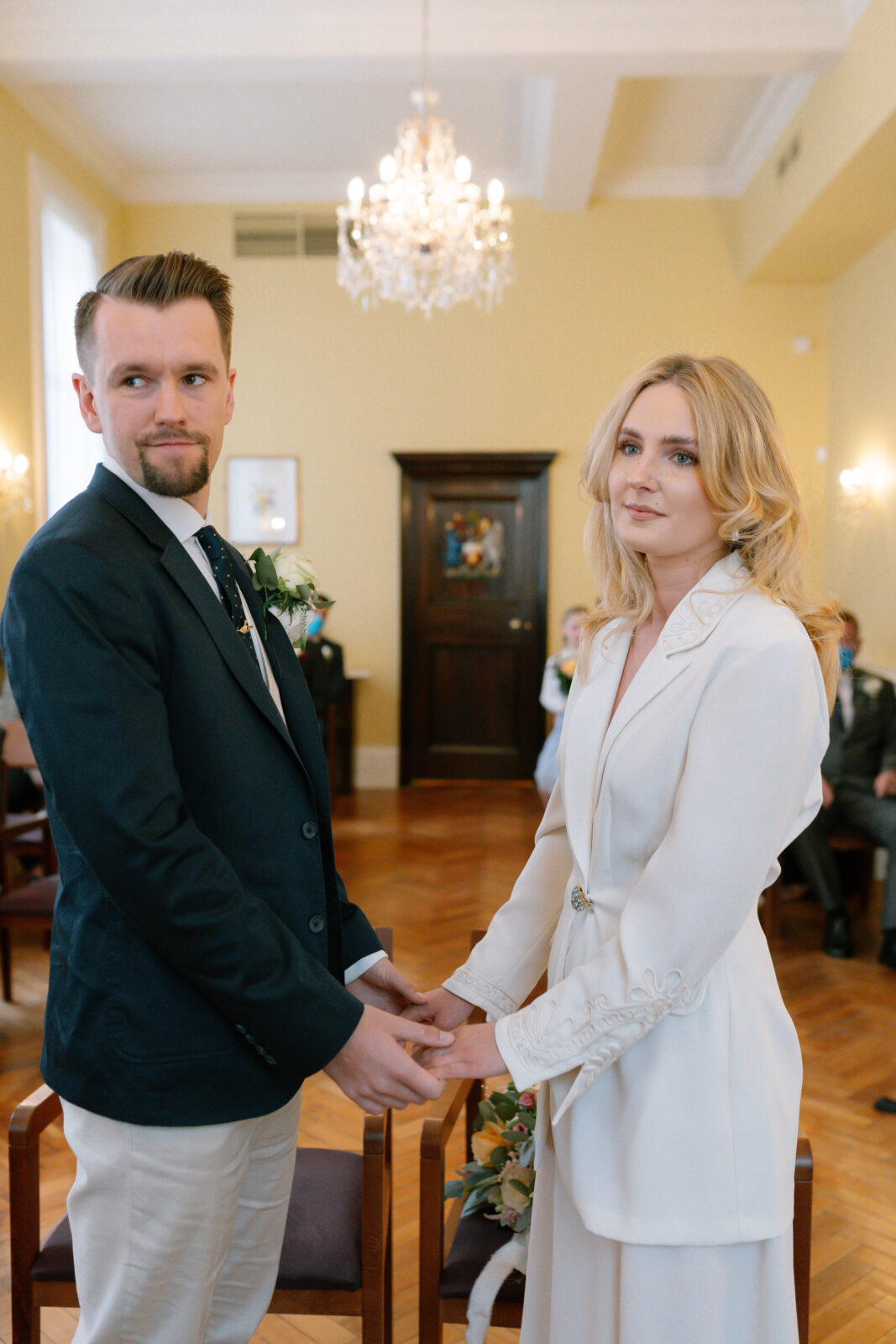 A couple dressed formally, holding hands and facing each other in a bright, elegant room with yellow walls, wooden floors, and a chandelier, suggesting a wedding or formal ceremony.