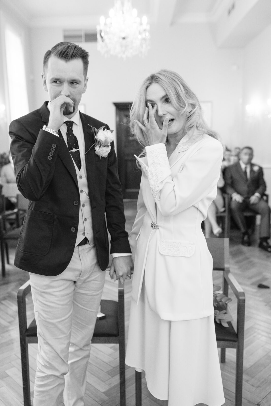 A couple dressed in formal wedding attire stand holding hands and smiling, playfully touching their noses with their fingers in a bright, elegant room with a chandelier.