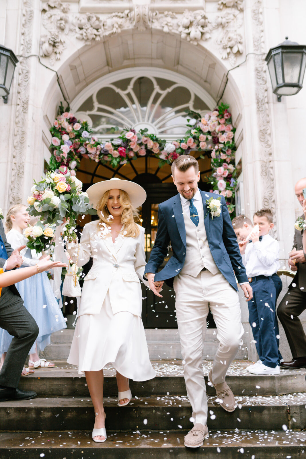 A newlywed couple, dressed in elegant light-colored outfits, walks down steps outside a beautifully decorated building as guests throw flower petals. The entrance is adorned with vibrant flowers. Both are smiling joyfully.
