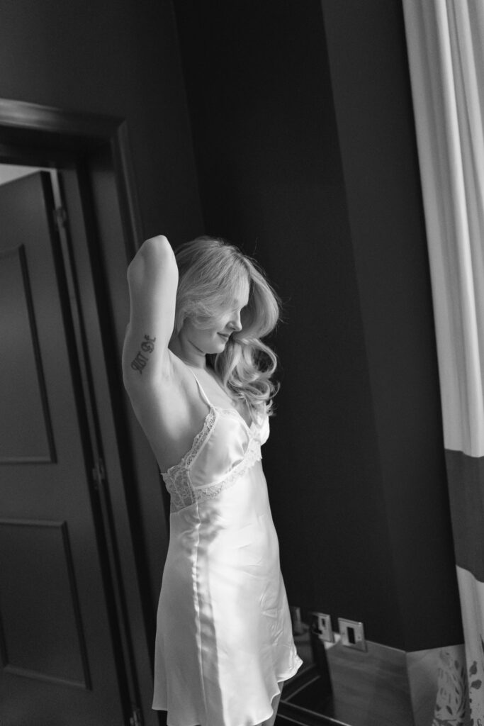 A woman in a satin slip dress stands near a window, smiling with her arms raised and her hair falling over her shoulders. The photo is in black and white.