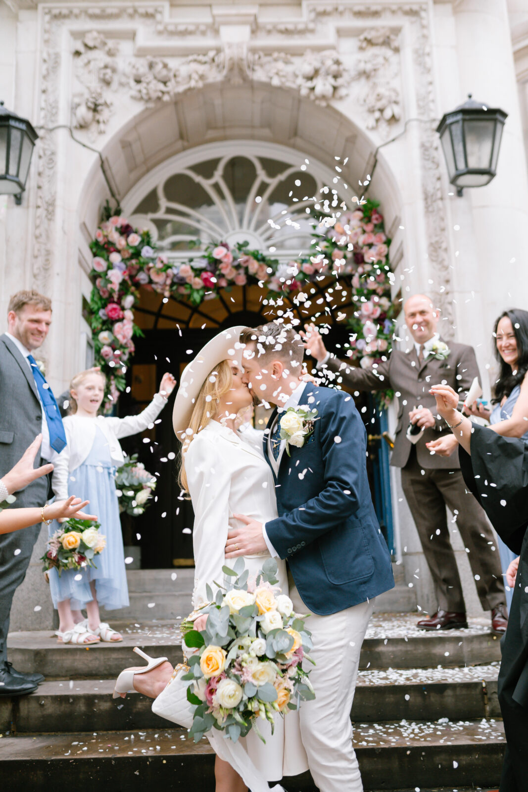 A newlywed couple kisses on the steps of a flower-adorned building while guests throw confetti. The bride holds a bouquet and wears a hat. Friends and family celebrate joyfully around them.
