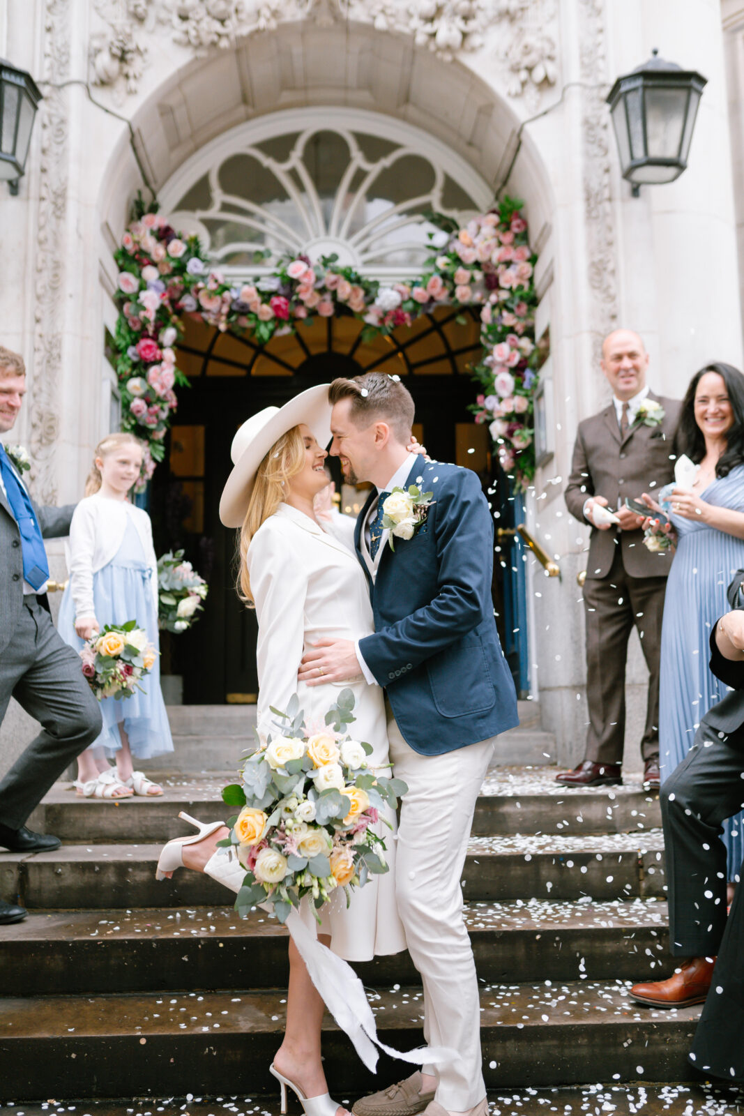 A newlywed couple embraces and smiles on the steps outside a building, surrounded by smiling guests throwing confetti. The bride holds a bouquet and wears a white dress and hat; floral arrangements decorate the entrance behind them.