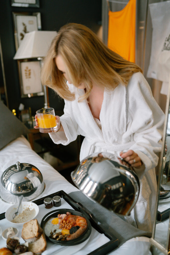 A woman in a white robe holds a glass of orange juice and lifts a metal food cover, revealing a breakfast tray with assorted dishes on a bed in a cozy, modern room.