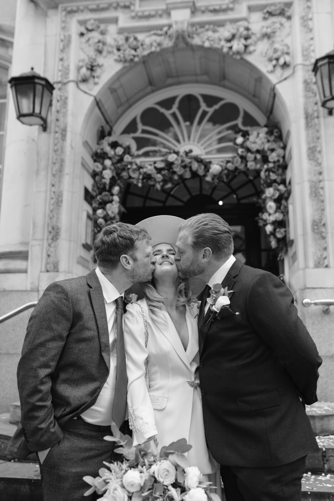 A bride smiles joyfully as two men in suits kiss her on each cheek in front of a building decorated with flowers and an arched doorway.