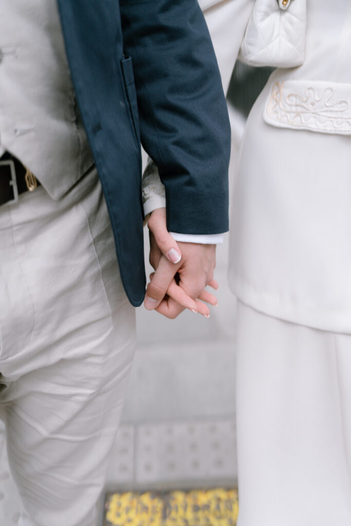A close-up of two people holding hands. One person is dressed in a light-colored suit, and the other is wearing a white outfit with decorative stitching. Their hands are gently intertwined.