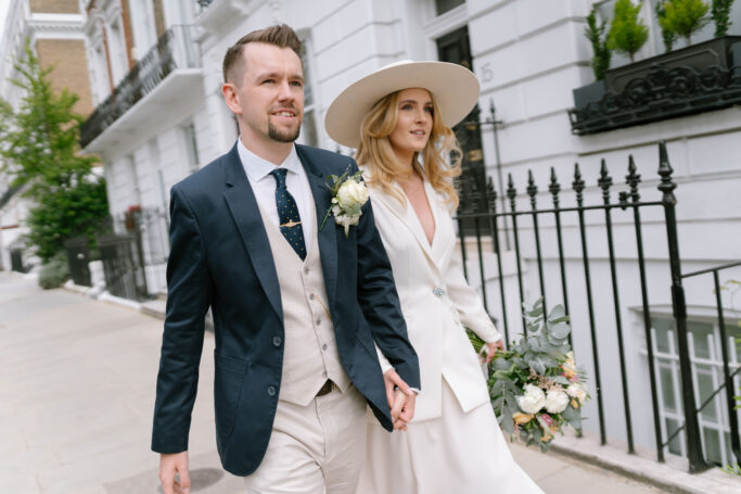 A couple dressed in elegant wedding attire walks hand in hand down a city sidewalk; the groom wears a navy suit, the bride a white dress and wide-brim hat, holding a bouquet of flowers.