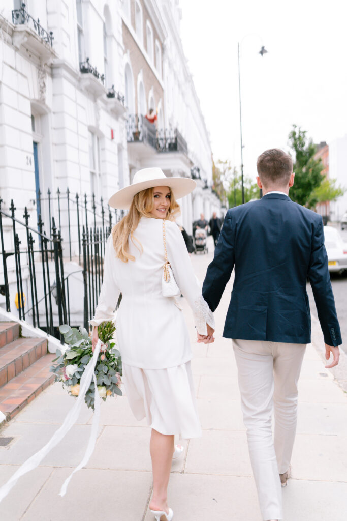 A woman in a white dress and wide-brimmed hat holds a bouquet and walks hand in hand with a man in a navy blazer and light pants down a city sidewalk, both smiling and looking happy.