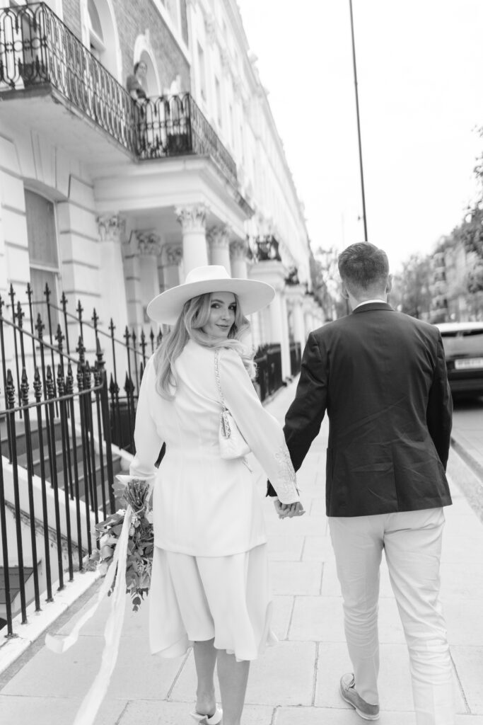 A woman in a white dress and wide-brimmed hat holds a bouquet and the hand of a man in a dark jacket as they walk down a city sidewalk, smiling back at the camera. The image is in black and white.