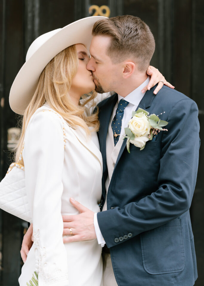 A couple dressed in wedding attire shares a kiss. The woman wears a white suit and wide-brimmed hat, holding a white bag, while the man wears a blue suit with a boutonnière and polka-dot tie.