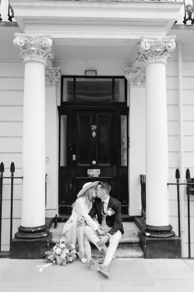 A couple dressed in wedding attire sits on the steps of a building, sharing a kiss. The building features two large columns, a black door with the number 29, and a bouquet lies on the ground beside them. The image is in black and white.