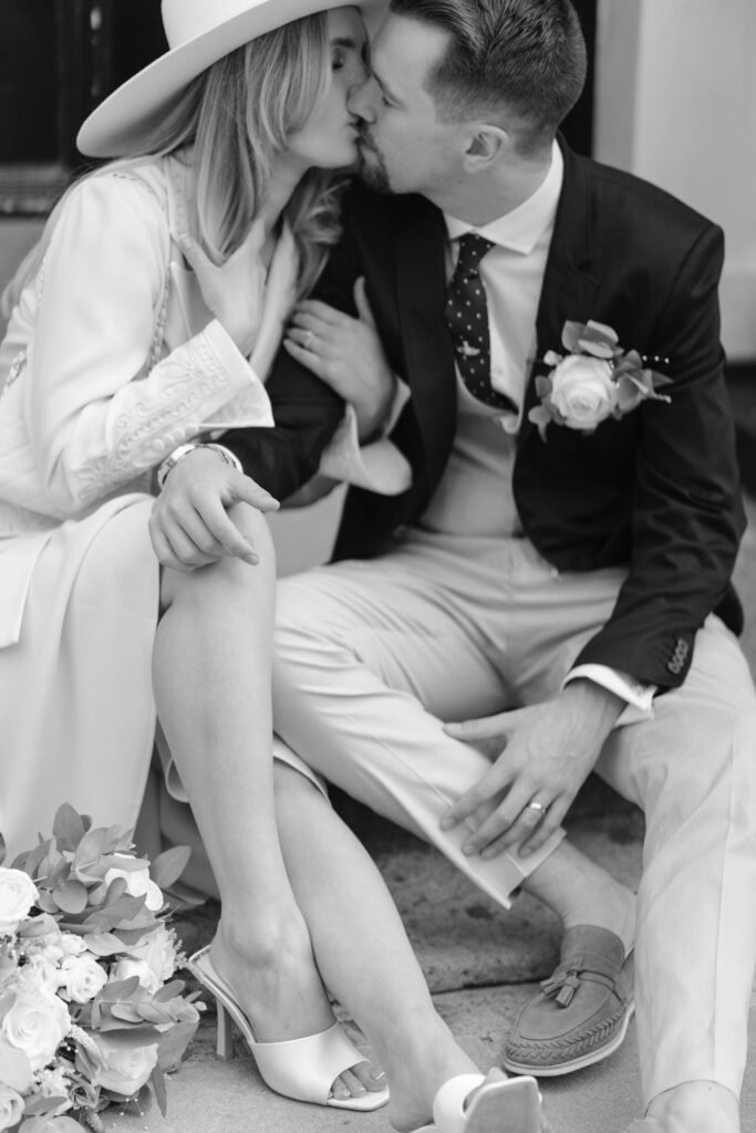 A bride and groom sit closely on stone steps, sharing a kiss. The bride wears a wide-brim hat and heeled sandals, while the groom wears a suit with a floral boutonniere. A bouquet rests near them. The image is in black and white.
