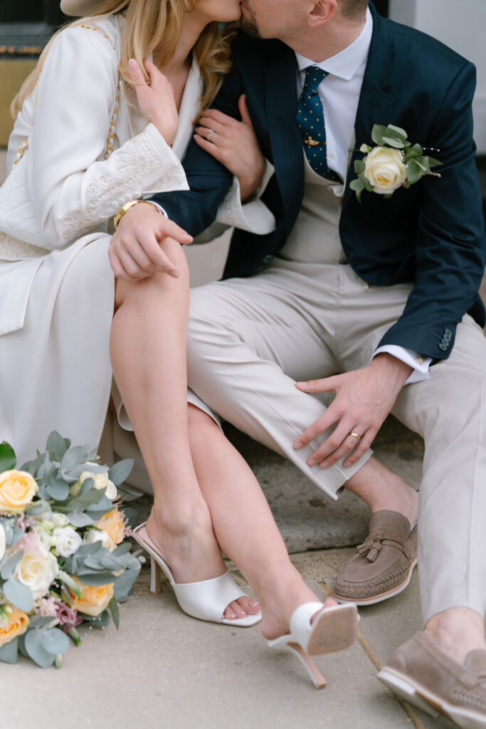 A couple dressed in elegant wedding attire sits closely on a step, sharing a kiss. The woman holds a bouquet of yellow and white flowers, and both wear rings, suggesting they are newlyweds.