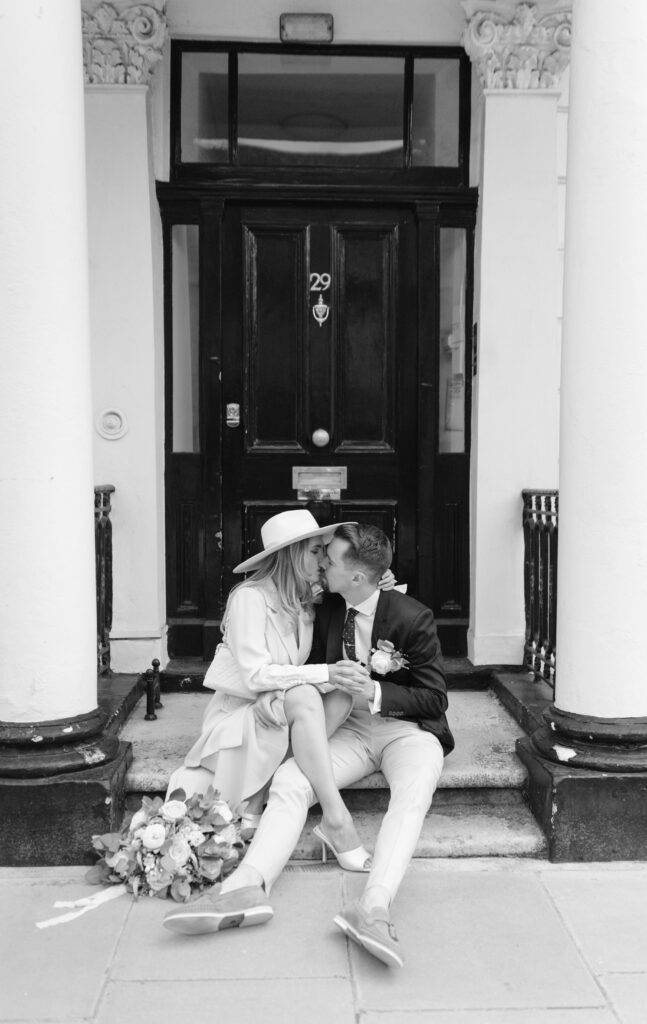 A couple in wedding attire sits on steps in front of a black door numbered 29. The woman wears a hat, the man a suit, and they share an intimate moment. A bouquet of flowers rests on the ground beside them.