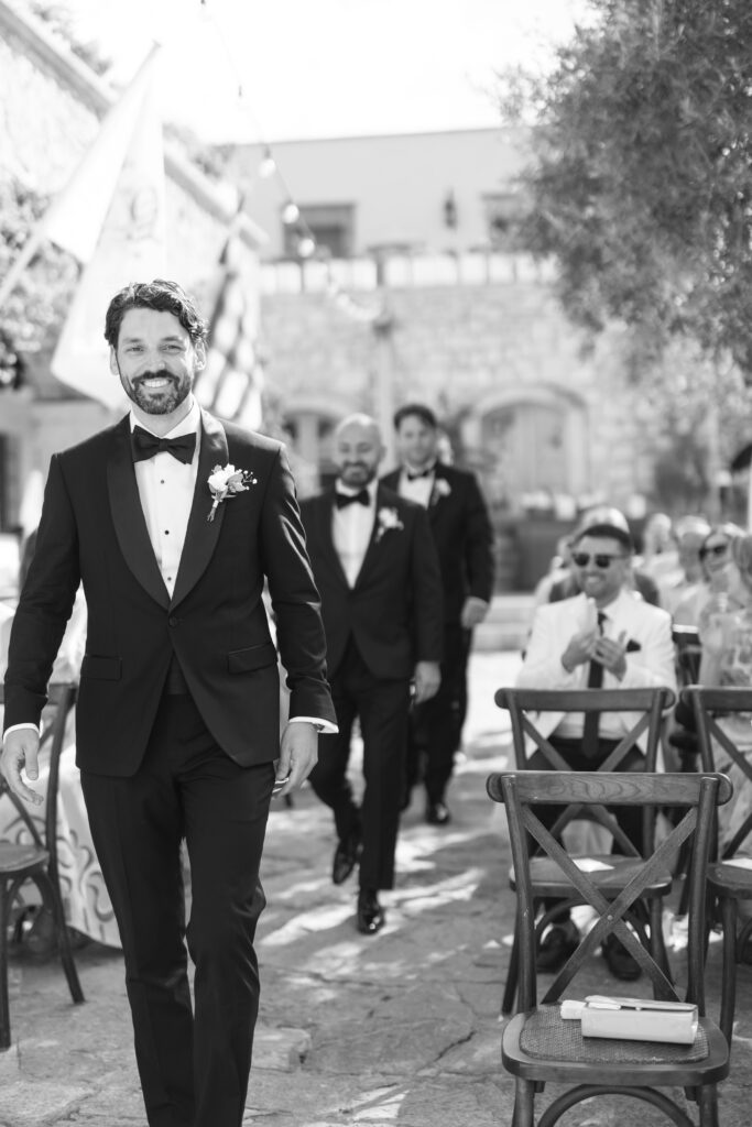 A man in a tuxedo walks down an outdoor aisle smiling, followed by other men in suits. Guests are seated and clapping in a bright, festive setting with stone buildings in the background.