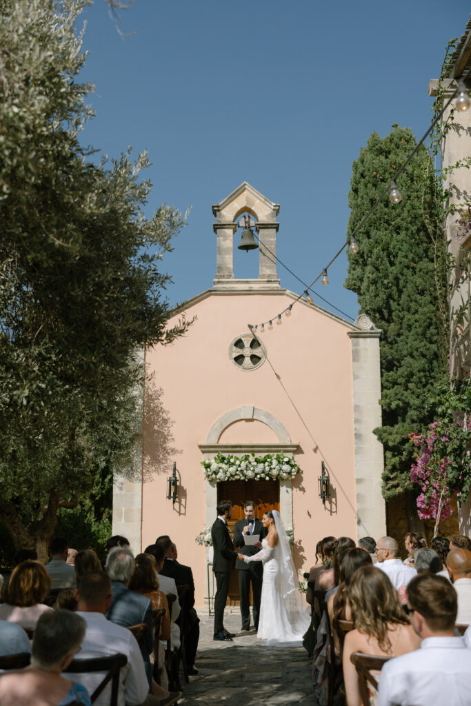 A bride and groom stand before an officiant outside a small, peach-colored chapel, surrounded by guests seated under a clear blue sky with string lights overhead.