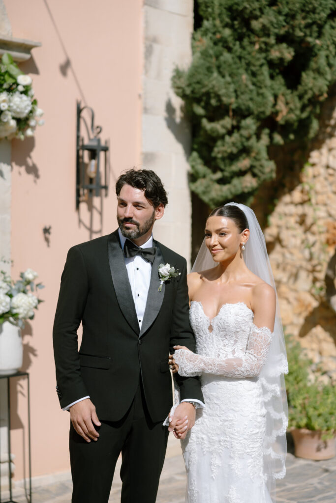 A bride in a white lace gown and veil stands arm-in-arm with a groom in a black tuxedo and bow tie, outdoors near a peach-colored wall decorated with flowers and greenery.