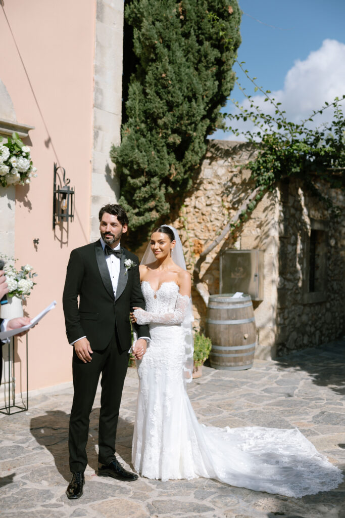 A bride in a white lace gown and veil stands beside a groom in a black tuxedo. They are outdoors on a stone patio, near a rustic building with greenery and flowers. Sunlight casts soft shadows across the scene.