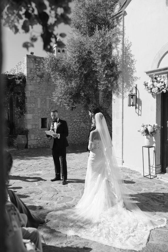 A bride in a wedding dress and veil stands outside facing a groom in a suit, who is reading from a piece of paper. They are in a stone courtyard next to a building with flowers by the door. The scene is in black and white.