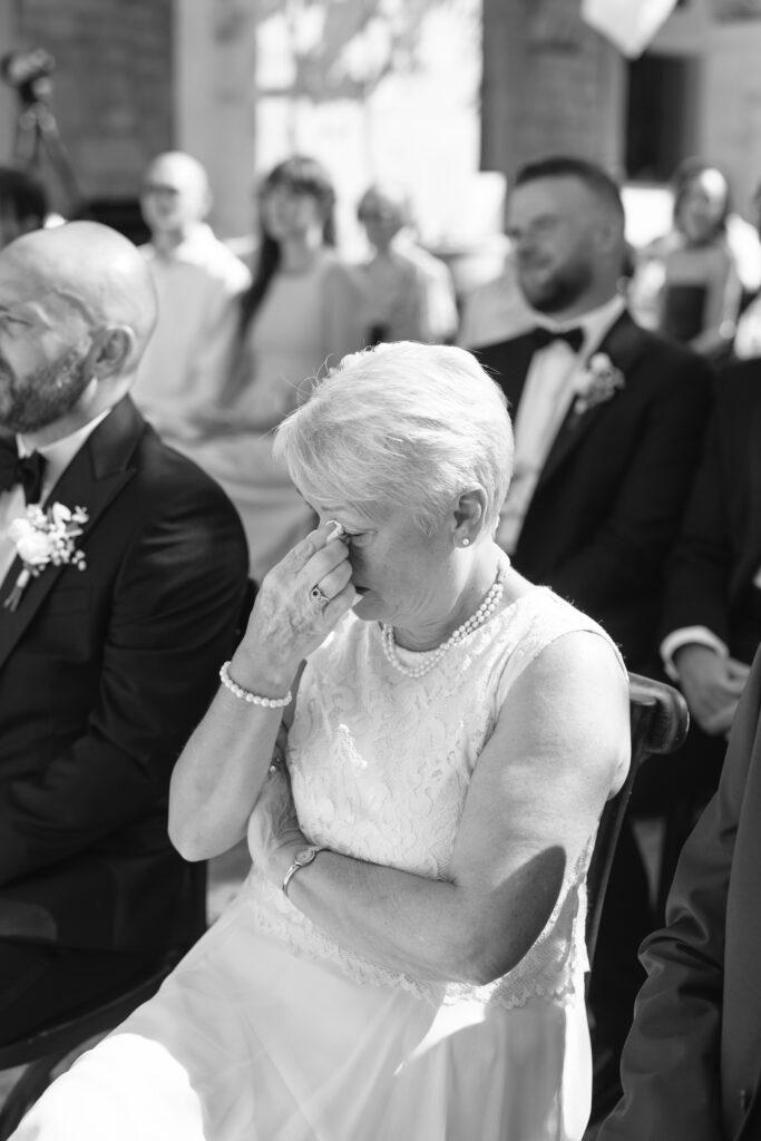 An older woman in a white dress sits among other guests at a formal event, wiping away tears with her hand, visibly emotional. The scene appears to be a wedding ceremony. The image is in black and white.