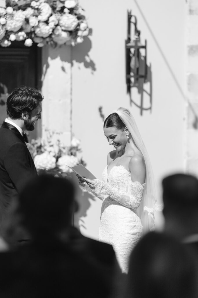 A bride in a lace wedding dress and veil reads vows to the groom during an outdoor ceremony, with floral decorations and guests visible in the foreground.