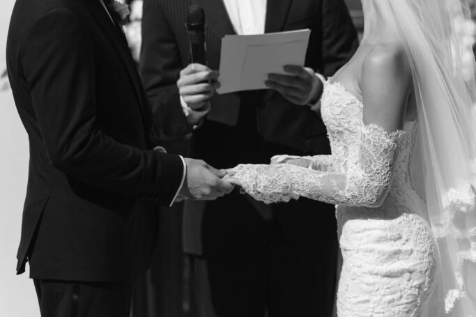 A bride and groom exchange rings during a wedding ceremony, with an officiant holding a microphone and speaking from notes. The bride wears a lace dress and veil; the groom is in a suit.