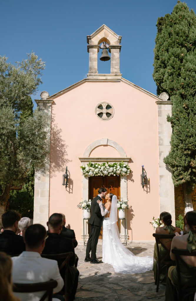 A bride and groom kiss in front of a pink stone chapel adorned with white flowers, surrounded by trees, as seated guests watch the outdoor wedding ceremony under a clear blue sky.