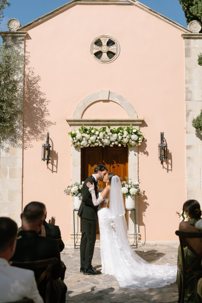 A bride and groom share a kiss in front of a small, peach-colored chapel adorned with white flowers, while guests seated outdoors watch and celebrate their wedding ceremony.