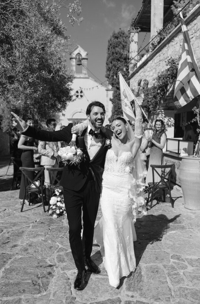 A joyful bride and groom walk arm in arm down a cobblestone path outside, surrounded by happy guests. The bride raises her bouquet and veil, both smiling widely, celebrating their wedding day.