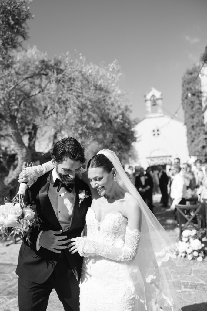 A beaming bride in a lace gown and veil embraces a smiling groom in a tuxedo outdoors, with guests and a chapel visible in the background under a sunny sky.