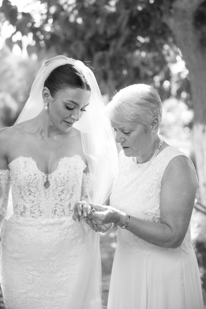A bride in a lace wedding gown and veil stands beside an older woman in a sleeveless dress. The older woman is holding the bride’s hand, examining or adjusting her ring outdoors.