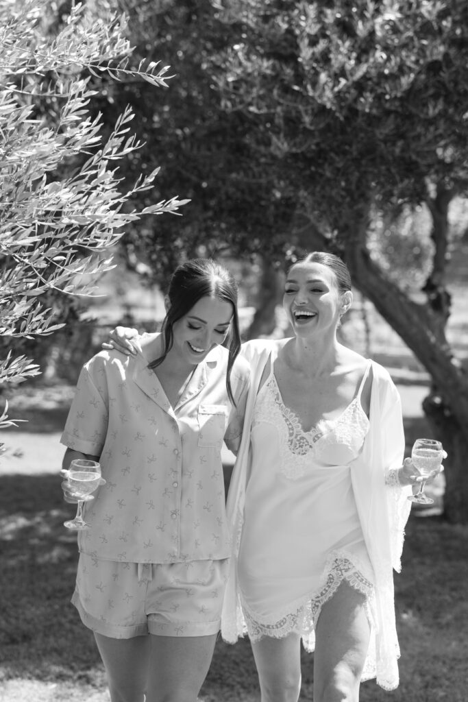 Two women in light sleepwear walk arm-in-arm outdoors, smiling and laughing, each holding a glass of wine. Sunlight filters through trees, creating a cheerful and relaxed atmosphere.