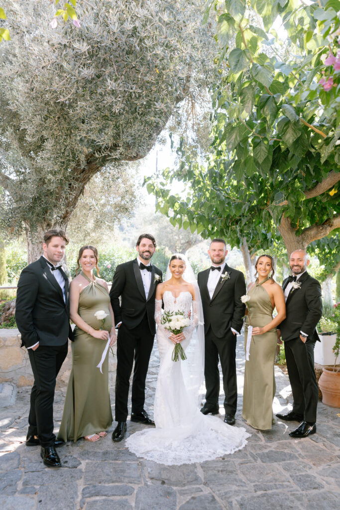 A bride and groom stand in the center, flanked by three groomsmen in black suits and two bridesmaids in sage green dresses, posing outdoors on a stone path beneath leafy trees.