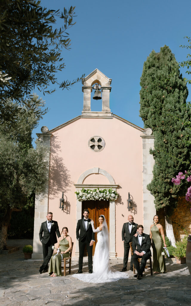 A bride and groom pose with four bridesmaids and groomsmen in front of a small, light pink stone chapel adorned with flowers, surrounded by trees, under a clear blue sky.