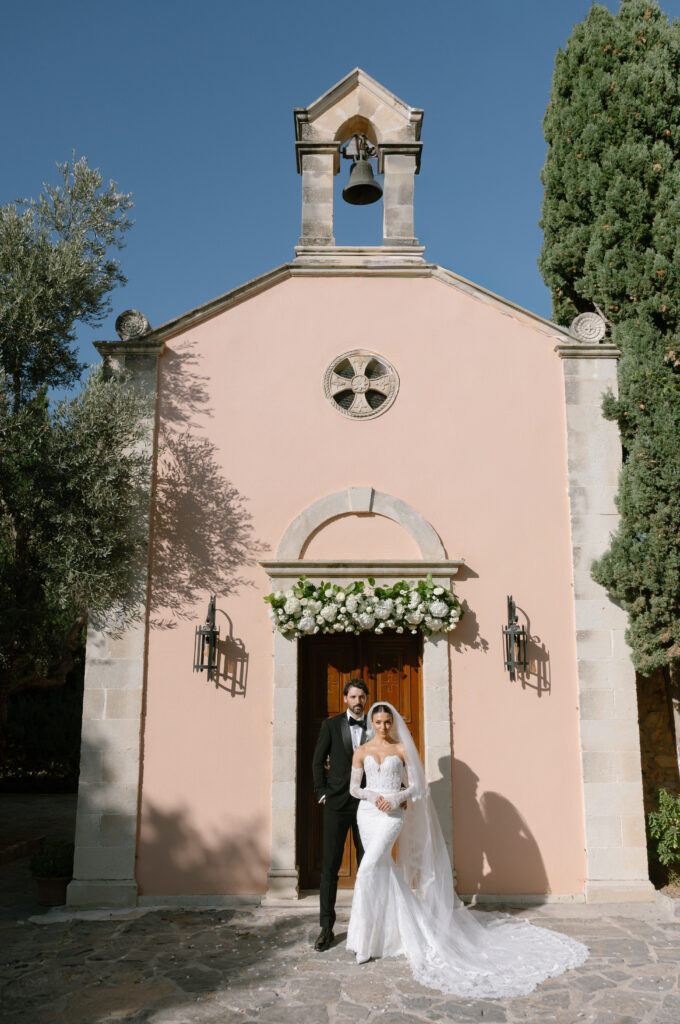 A bride and groom stand in front of a small, peach-colored chapel with floral decorations above the door, on a sunny day. The bride wears a long white gown and veil; the groom wears a black suit and bow tie.