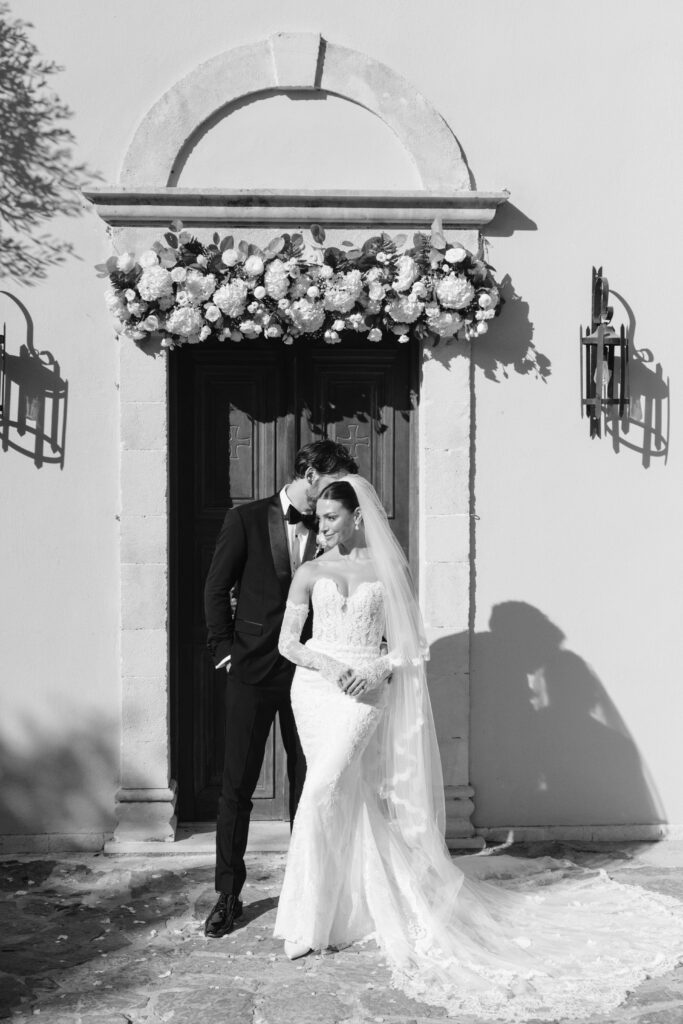 A bride and groom stand outside a stone building’s arched doorway adorned with flowers. The groom in a suit gently kisses the bride’s head; she wears a long lace gown and veil, both smiling in the sunlight. Black and white photo.