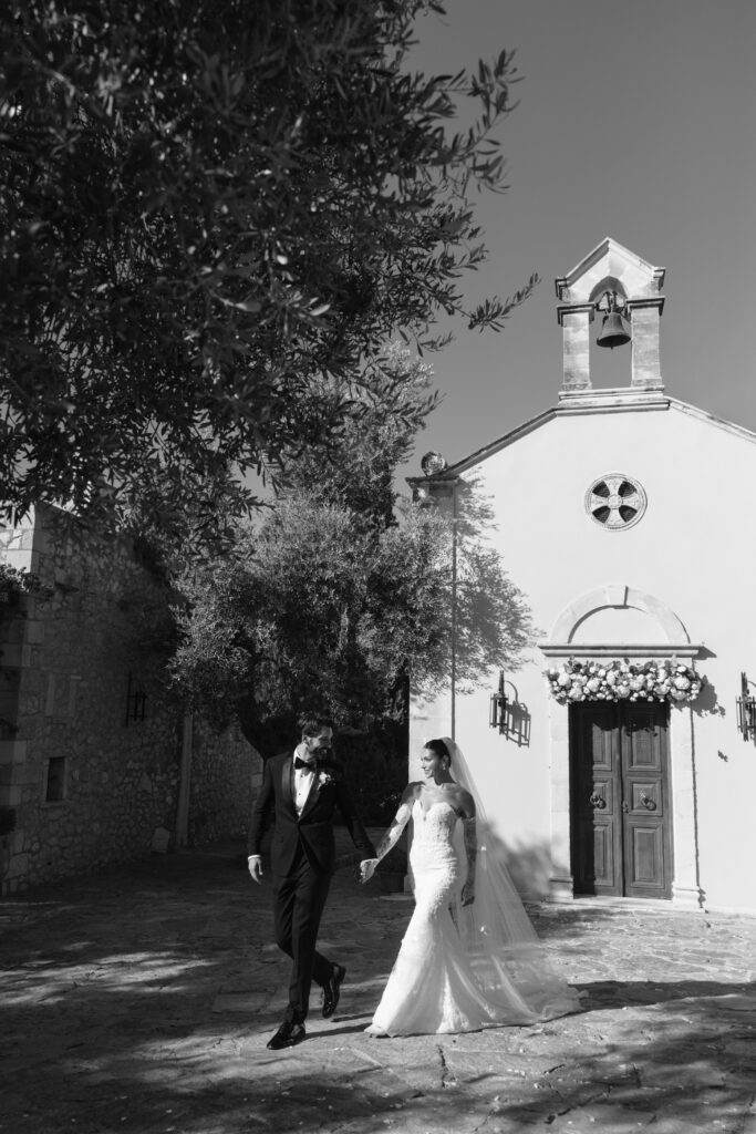 A bride and groom, dressed in wedding attire, hold hands and walk outside a small, decorated chapel under a tree on a sunny day. The scene is in black and white.