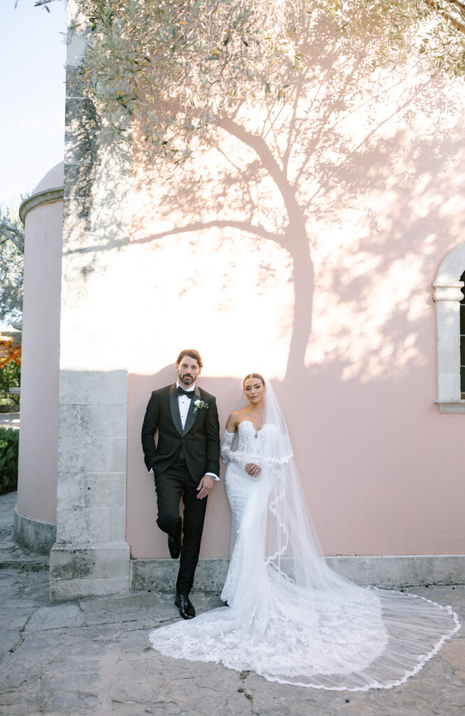 A bride in a long white gown and veil stands beside a groom in a black tuxedo, both posing against a sunlit pink wall with a tree shadow, near a stone building and window.