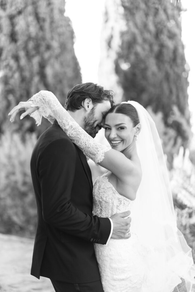A joyful bride in a lace wedding dress and veil embraces a smiling groom in a suit outdoors. The bride’s arm is wrapped around the groom and she looks at the camera, while the groom faces her with trees in the blurred background.