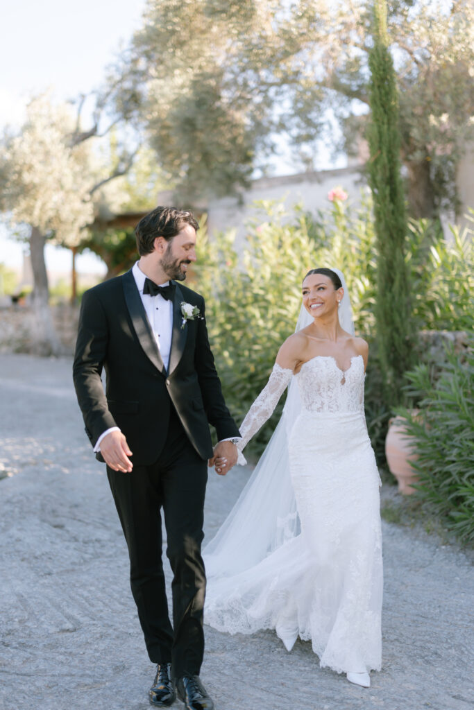 A bride in a lace, off-the-shoulder wedding dress and a groom in a black tuxedo walk hand-in-hand outdoors, smiling at each other. Lush greenery and trees fill the bright, sunlit background.
