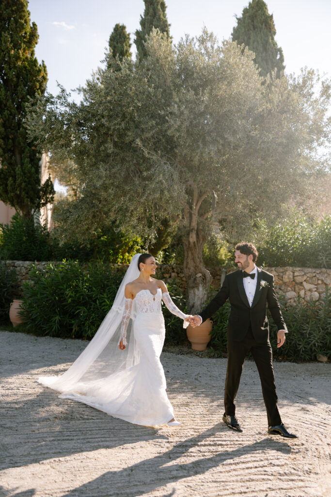A bride in a long white dress and veil holds hands with a groom in a black tuxedo as they walk outdoors on a sunny day, with trees and greenery in the background.