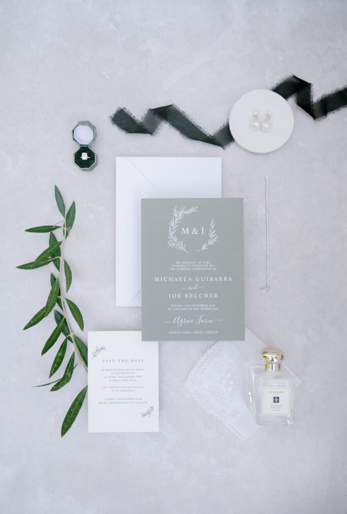 Flat lay of elegant wedding invitation suite with green and white cards, an envelope, a diamond ring in a box, pearl earrings, a necklace, black ribbon, greenery sprig, lace, and a Jo Malone perfume bottle.