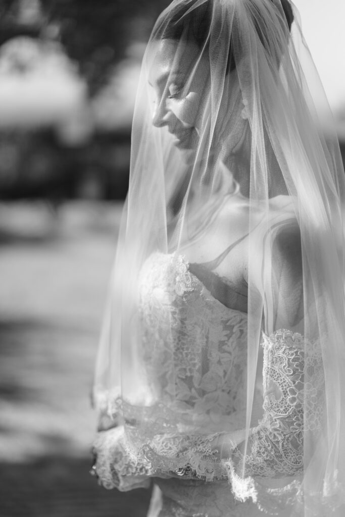 A bride in a lace wedding dress and veil smiles softly, standing outdoors in natural light. The photo is black and white, giving it a timeless, elegant feel.