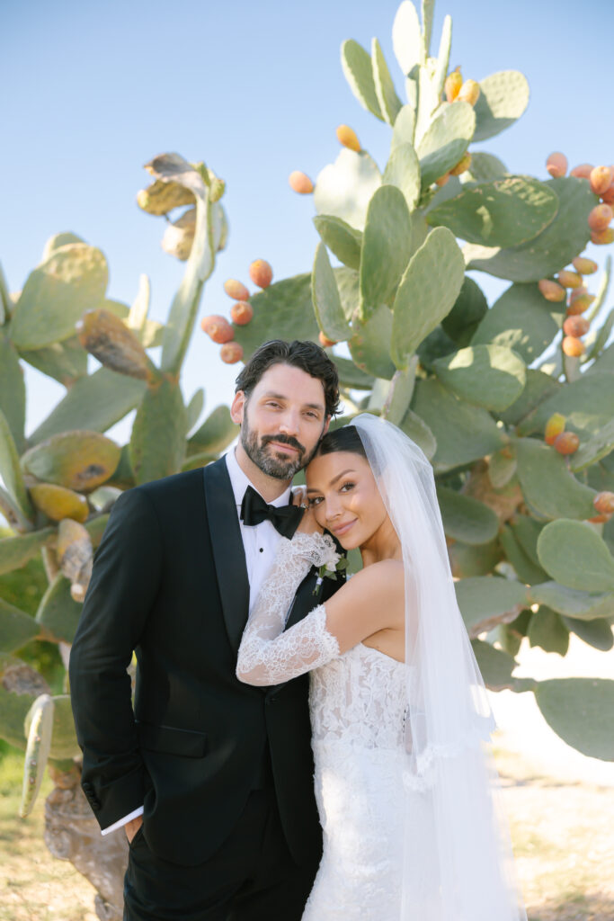 A bride in a lace wedding dress and veil leans her head on the groom’s shoulder. The groom is dressed in a black tuxedo. They stand in front of a large cactus with fruit under a clear blue sky.