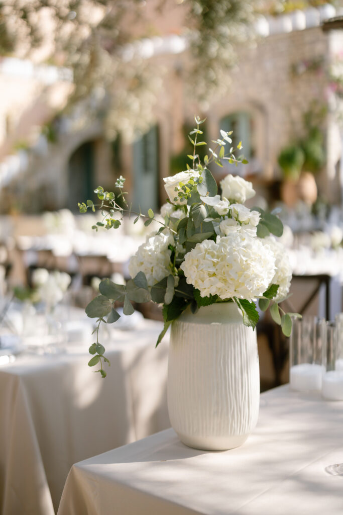 A white ceramic vase filled with white hydrangeas, roses, and green foliage sits on a table with a cream tablecloth in an outdoor setting, with blurred tables and stone buildings in the background.