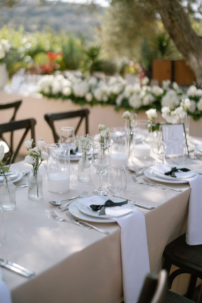 Elegant outdoor table setting with white plates, glassware, white napkins, and small vases of white flowers on a beige tablecloth. Lush greenery and white floral arrangements are visible in the background.