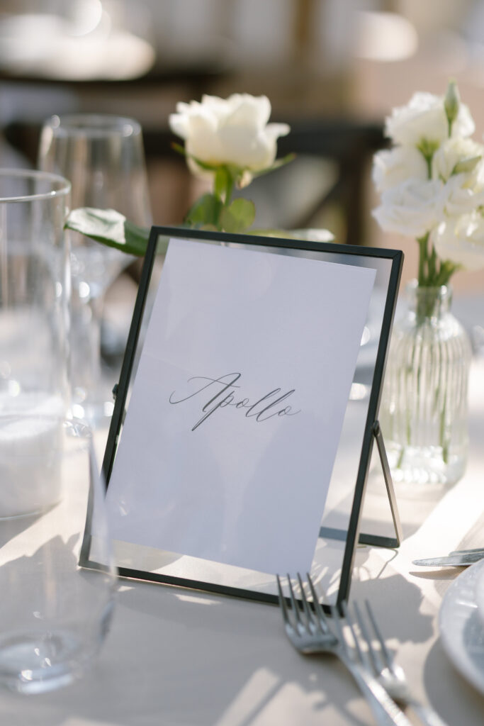 A table setting with elegant glassware and white flowers, featuring a framed card labeled Apollo in cursive script, suggesting a table name or seating arrangement at an event.