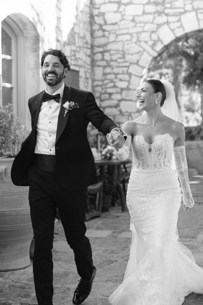 A bride and groom, smiling and holding hands, walk outdoors at a stone courtyard. The bride wears a strapless lace gown and veil, the groom wears a tuxedo with a bow tie and boutonniere.